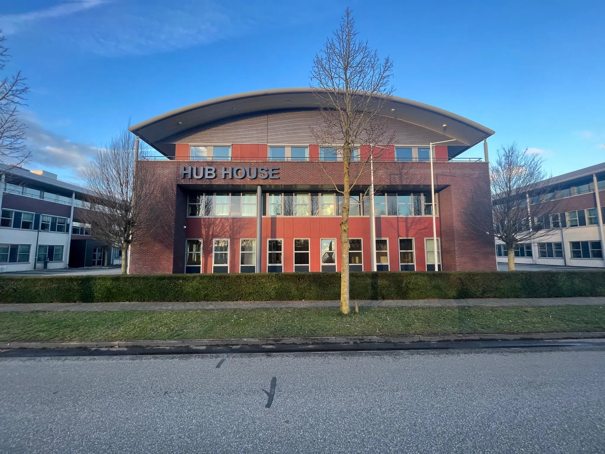 Voorzijde van het moderne kantoorgebouw "HUB HOUSE" met roodbruine gevel en omliggend groen.